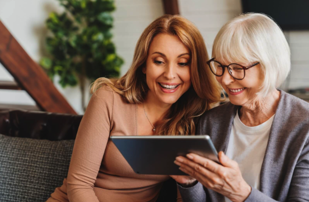 Two women, one younger and one older, smiling and looking at a tablet together during an assisted living visitation.