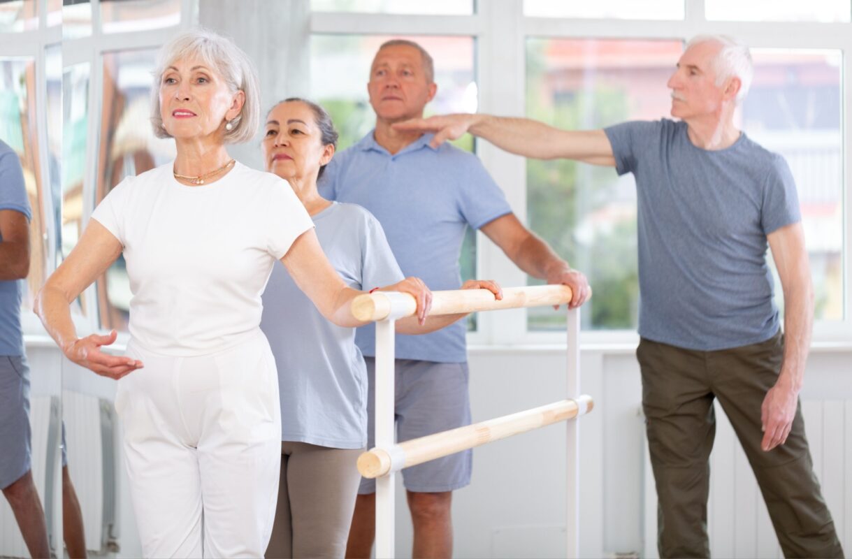 Four seniors participate in a bright, mirrored studio, standing at a barre and following posture exercises—one of the best exercises to support flexibility and balance.