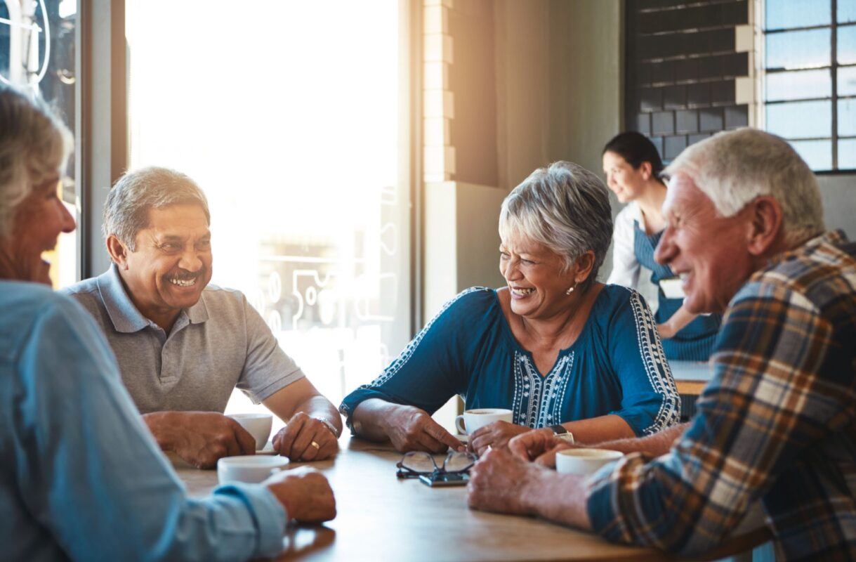 Four older adults sit around a table in a bright café, smiling and talking over cups of coffee, with another person in the background.