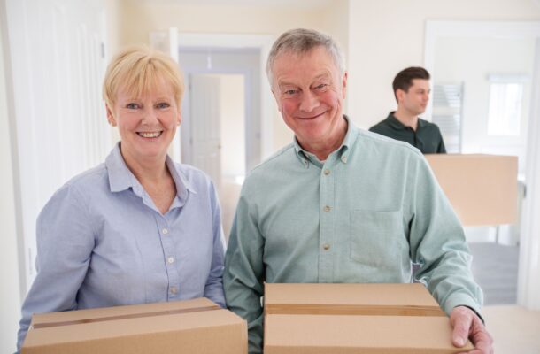 Two older adults holding cardboard boxes stand in the foreground, smiling, while a younger person carries a box in the background inside a bright room.